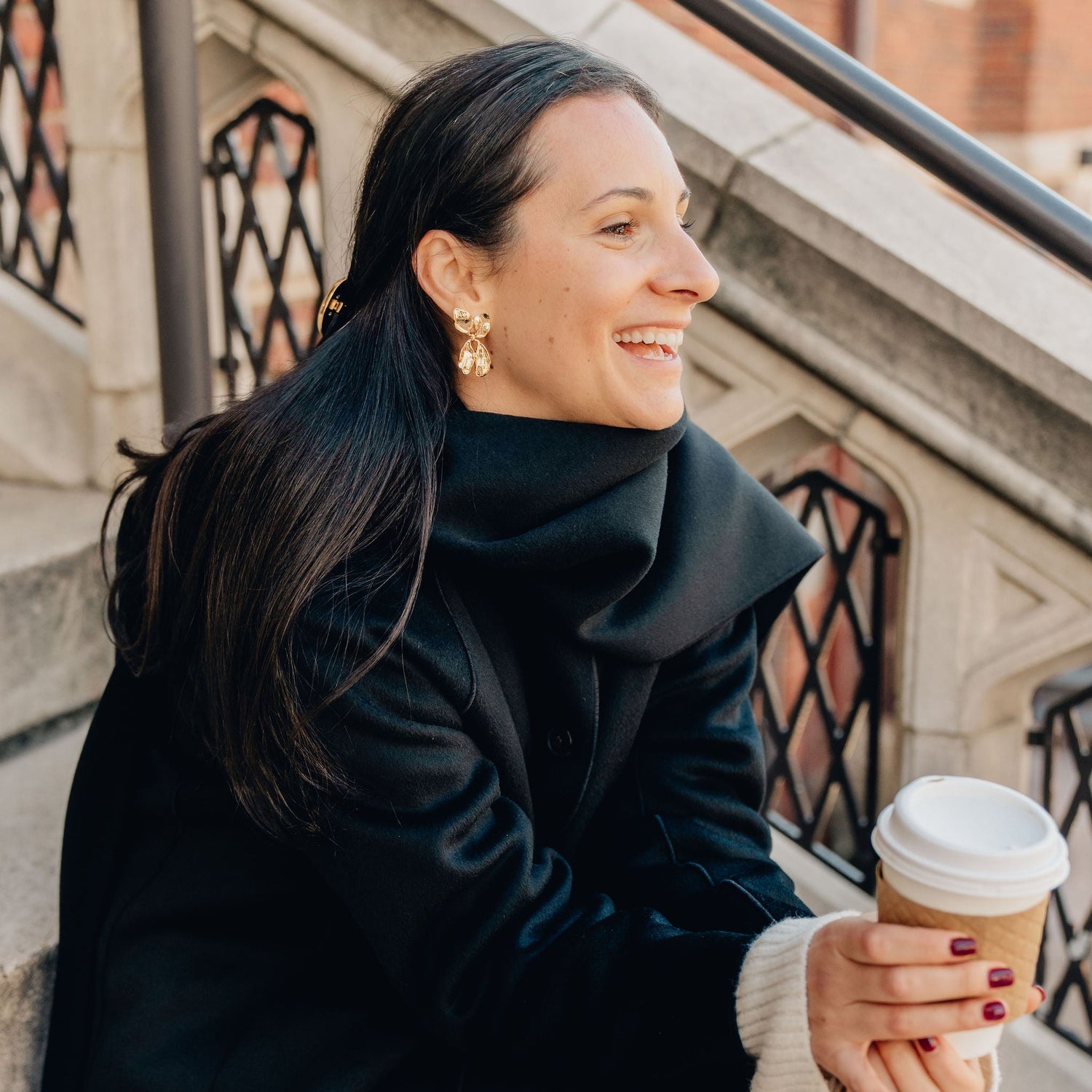 Woman in a black coat holding a coffee cup outdoors wearing statement holiday earrings.