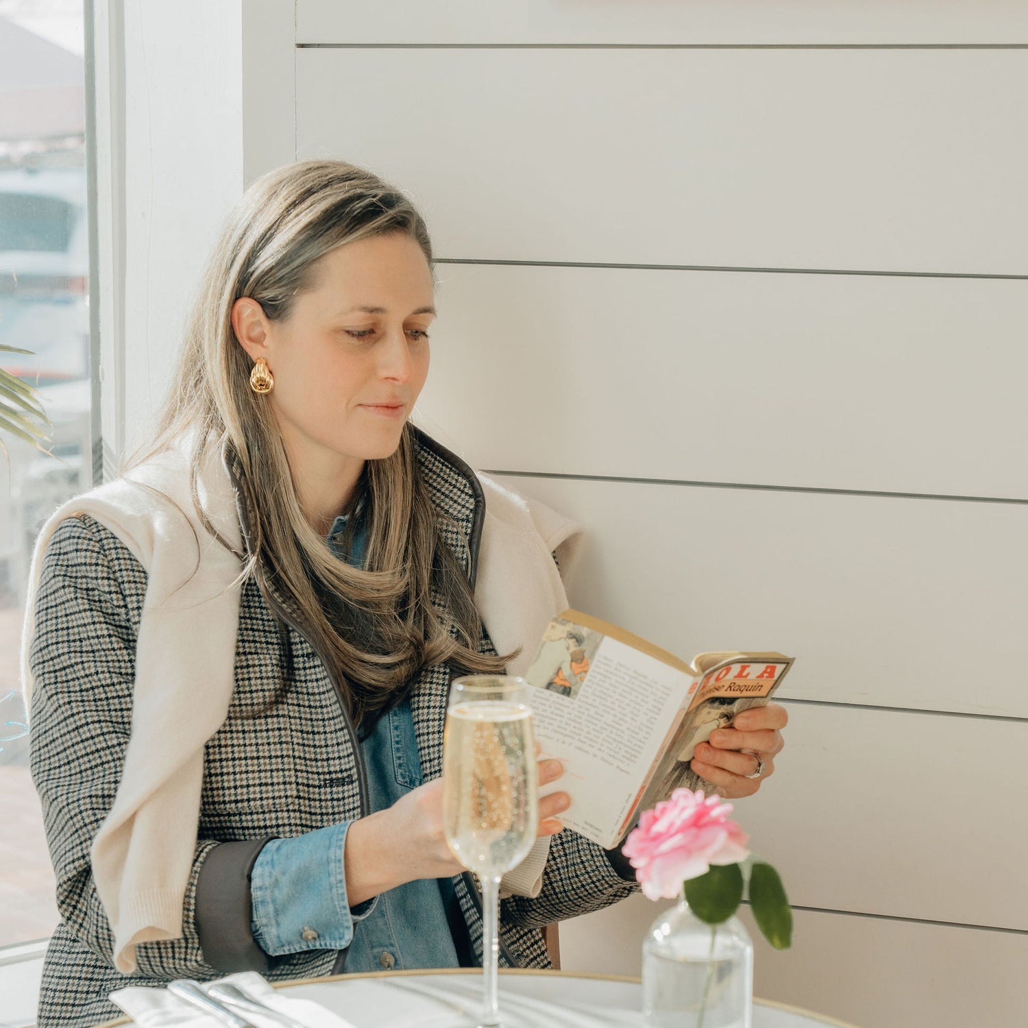 Woman sitting at a table with a glass of wine and a book, surrounded by utensils and a vase with flowers wearing statement classic holiday earrings