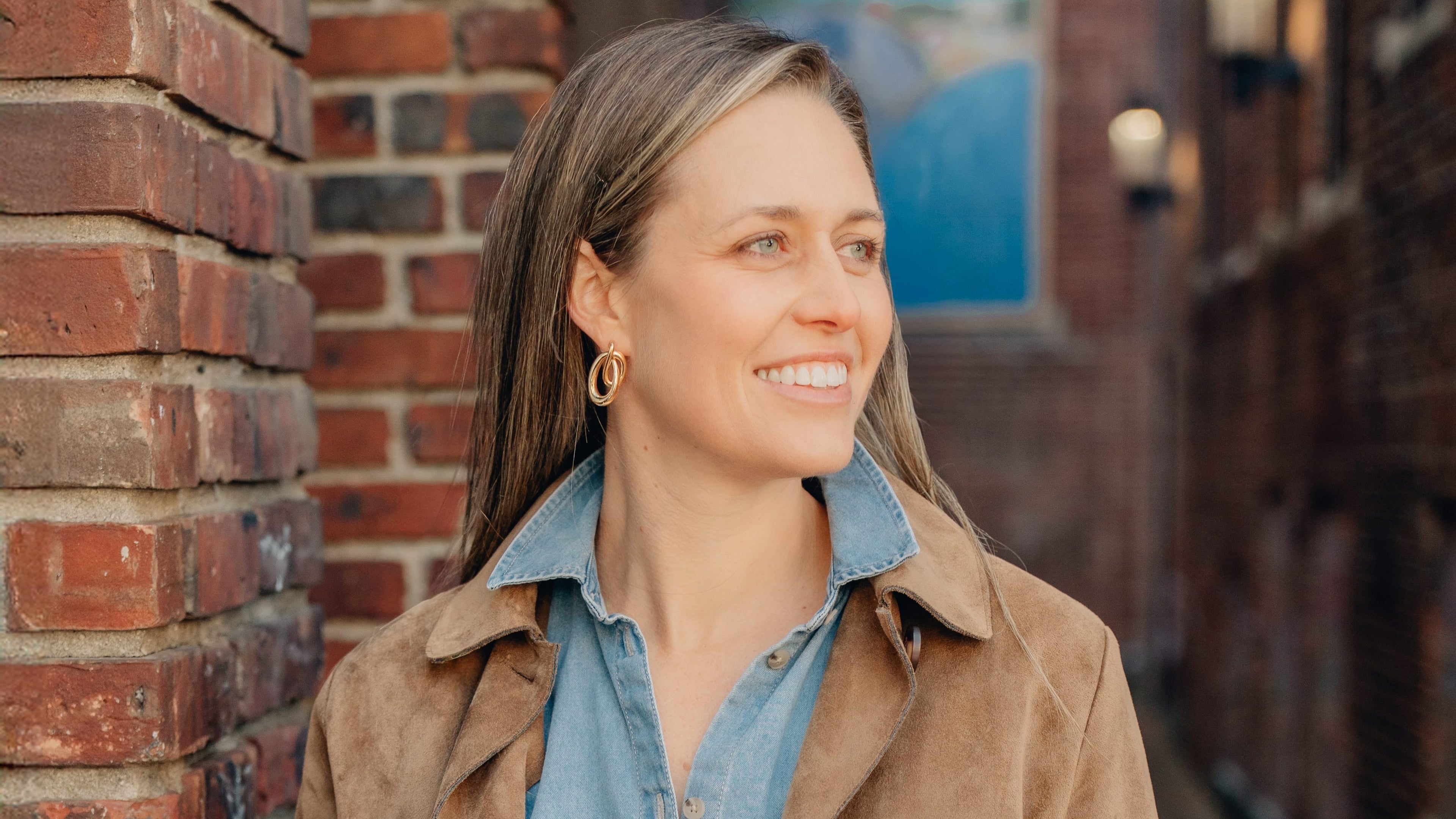 Woman standing against a brick wall wearing a brown coat, blue shirt, and double loop earrings..