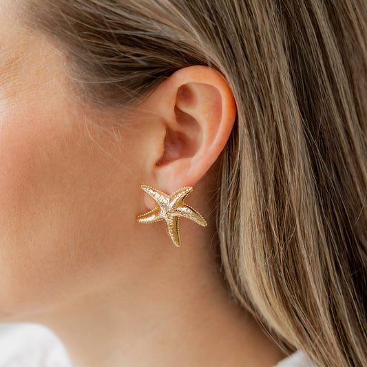 Gold starfish earring worn by a person with light skin and blonde hair, wearing a white shirt.